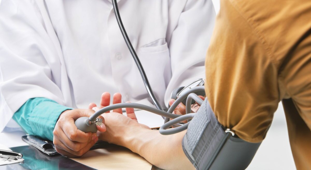 Das Bild zeigt eine medizinische Untersuchung, bei der ein Arzt oder eine medizinische Fachkraft gerade dabei ist, den Blutdruck einer Person zu messen. Die Person mit dem Blutdruckmessgerät um den Arm sitzt, während der Arzt die Manschette des Geräts aufpumpt und durch das Stethoskop hört. Auf dem Tisch liegt ein Tablet oder eine Patientenakte. Der Fokus liegt auf den Händen, dem Blutdruckmessgerät und dem Stethoskop, während die Identitäten der Personen anonym bleiben und die Szene eine professionelle medizinische Atmosphäre vermittelt.