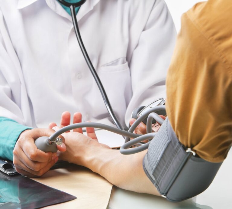 Das Bild zeigt eine medizinische Untersuchung, bei der ein Arzt oder eine medizinische Fachkraft gerade dabei ist, den Blutdruck einer Person zu messen. Die Person mit dem Blutdruckmessgerät um den Arm sitzt, während der Arzt die Manschette des Geräts aufpumpt und durch das Stethoskop hört. Auf dem Tisch liegt ein Tablet oder eine Patientenakte. Der Fokus liegt auf den Händen, dem Blutdruckmessgerät und dem Stethoskop, während die Identitäten der Personen anonym bleiben und die Szene eine professionelle medizinische Atmosphäre vermittelt. | © imago/Design Pics