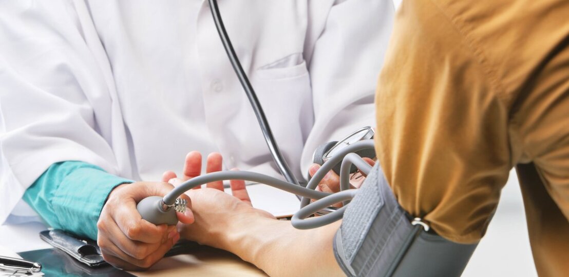 Das Bild zeigt eine medizinische Untersuchung, bei der ein Arzt oder eine medizinische Fachkraft gerade dabei ist, den Blutdruck einer Person zu messen. Die Person mit dem Blutdruckmessgerät um den Arm sitzt, während der Arzt die Manschette des Geräts aufpumpt und durch das Stethoskop hört. Auf dem Tisch liegt ein Tablet oder eine Patientenakte. Der Fokus liegt auf den Händen, dem Blutdruckmessgerät und dem Stethoskop, während die Identitäten der Personen anonym bleiben und die Szene eine professionelle medizinische Atmosphäre vermittelt. | © imago/Design Pics