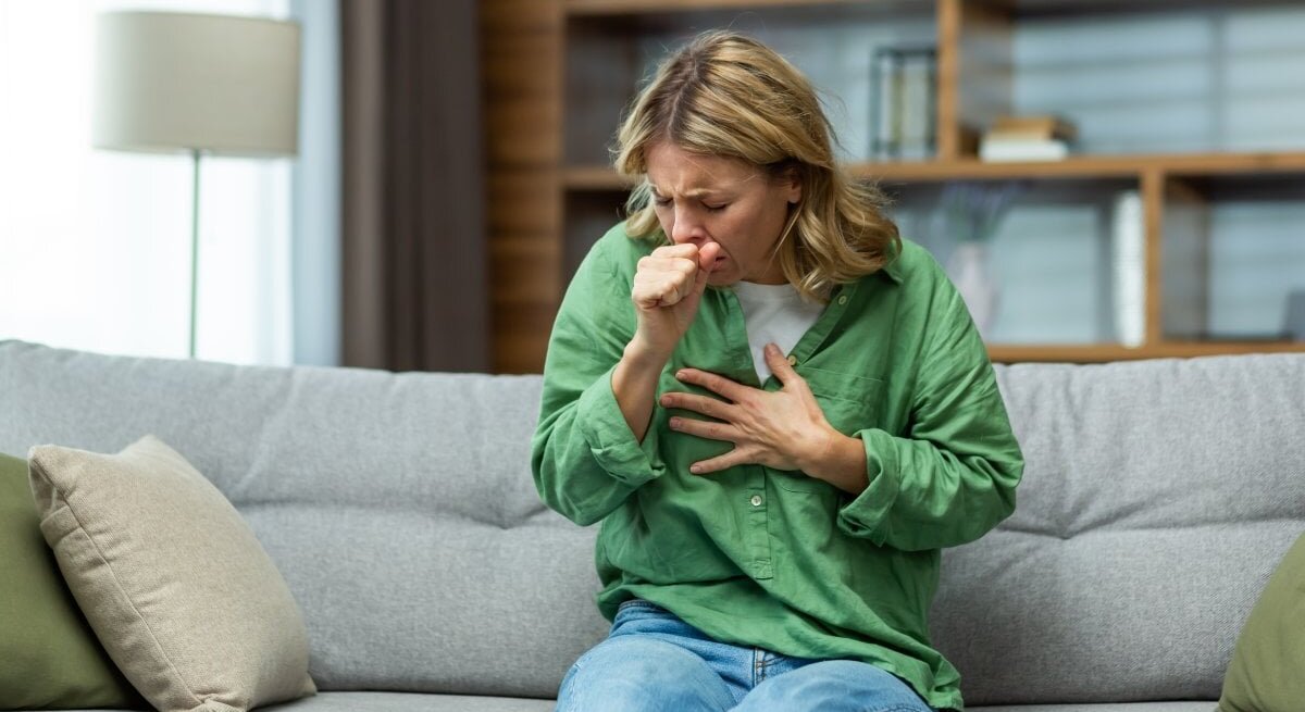 Das Bild zeigt eine Frau mittleren Alters, die auf einem Sofa sitzt und sich mit einer Hand auf die Brust fasst, während sie mit der anderen Hand ihren Mund bedeckt, als würde sie husten. Sie wirkt besorgt oder unwohl, möglicherweise aufgrund des Reizhustens oder verschleimten Hustens. Die Umgebung deutet auf ein häusliches Wohnzimmer hin, mit einem modernen, ruhigen Hintergrund, der das Hauptaugenmerk auf die Frau lenkt. Ihre Kleidung ist leger, mit einem grünen Hemd und blauen Jeans, was darauf hindeutet, dass sie sich in einer entspannten Umgebung befindet.