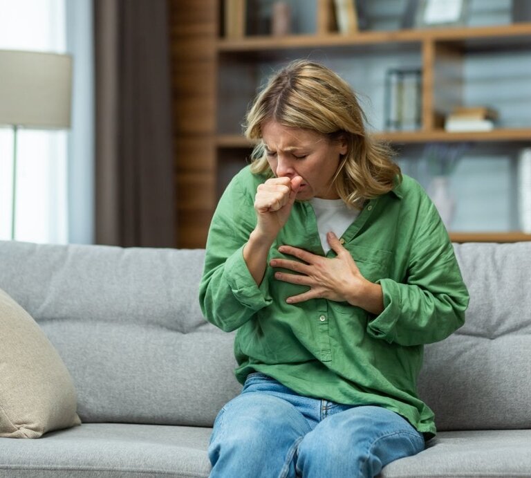 Das Bild zeigt eine Frau mittleren Alters, die auf einem Sofa sitzt und sich mit einer Hand auf die Brust fasst, während sie mit der anderen Hand ihren Mund bedeckt, als würde sie husten. Sie wirkt besorgt oder unwohl, möglicherweise aufgrund des Reizhustens oder verschleimten Hustens. Die Umgebung deutet auf ein häusliches Wohnzimmer hin, mit einem modernen, ruhigen Hintergrund, der das Hauptaugenmerk auf die Frau lenkt. Ihre Kleidung ist leger, mit einem grünen Hemd und blauen Jeans, was darauf hindeutet, dass sie sich in einer entspannten Umgebung befindet. | © shutterstock/ voronaman