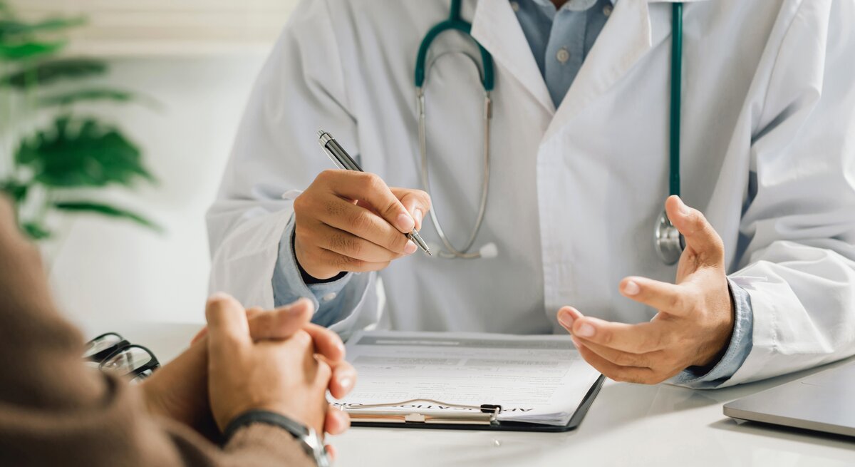 Ein Arzt spricht mit Patient. Gesichter sind nicht zu sehen, dafür gestikulierende Hände und ein Klippboard.