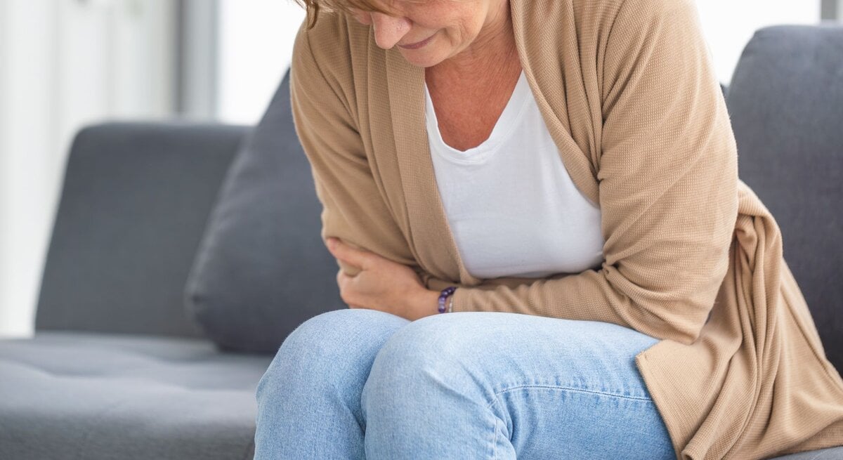 Eine Frau hält sich vor Schmerzen den Bauch und sitzt dabei auf einem grauen Sofa.