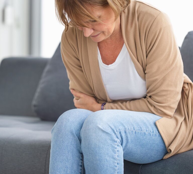 Eine Frau hält sich vor Schmerzen den Bauch und sitzt dabei auf einem grauen Sofa. | © Shutterstock/JU.STOCKER