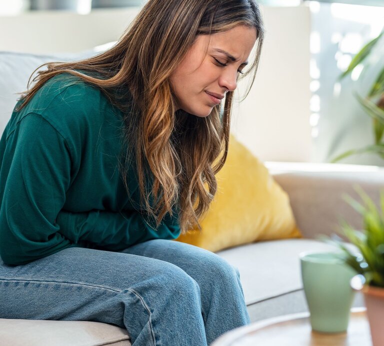 Eine Frau, die einen dunkelgrünen Pullover trägt und braune Haare hat, sitzt auf einem hellen Sofa und hält sich mit schmerzverzerrtem Gesicht den linken Bauchbereich. Sie scheint starke Schmerzen zu haben. | © Adobe Stock/ nenetus