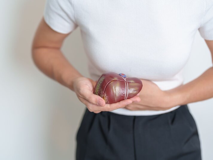 Junge Frau in weißem Shirt hält ein Lebermodell und fasst sich mit schmerzverzerrtem Gesicht an den rechten Oberbauch. | © Adobe Stock / Jo Panuwat D