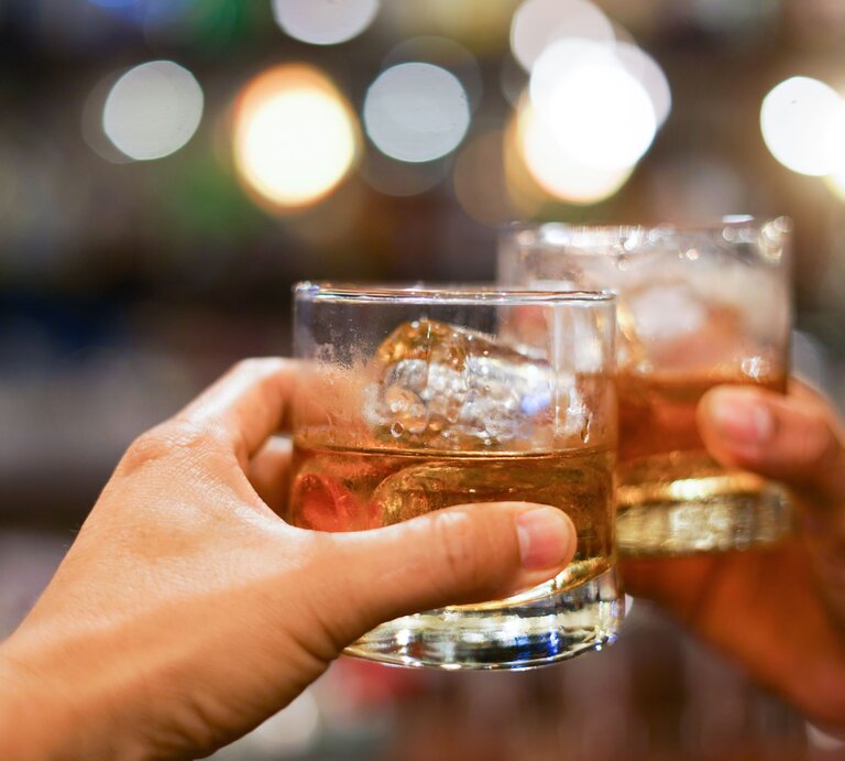 Zwei Hände stoßen mit gefüllten Whiskygläsern vor verschwommenem, buntem Hintergrund an. | © Getty Images/krisanapong detraphiphat