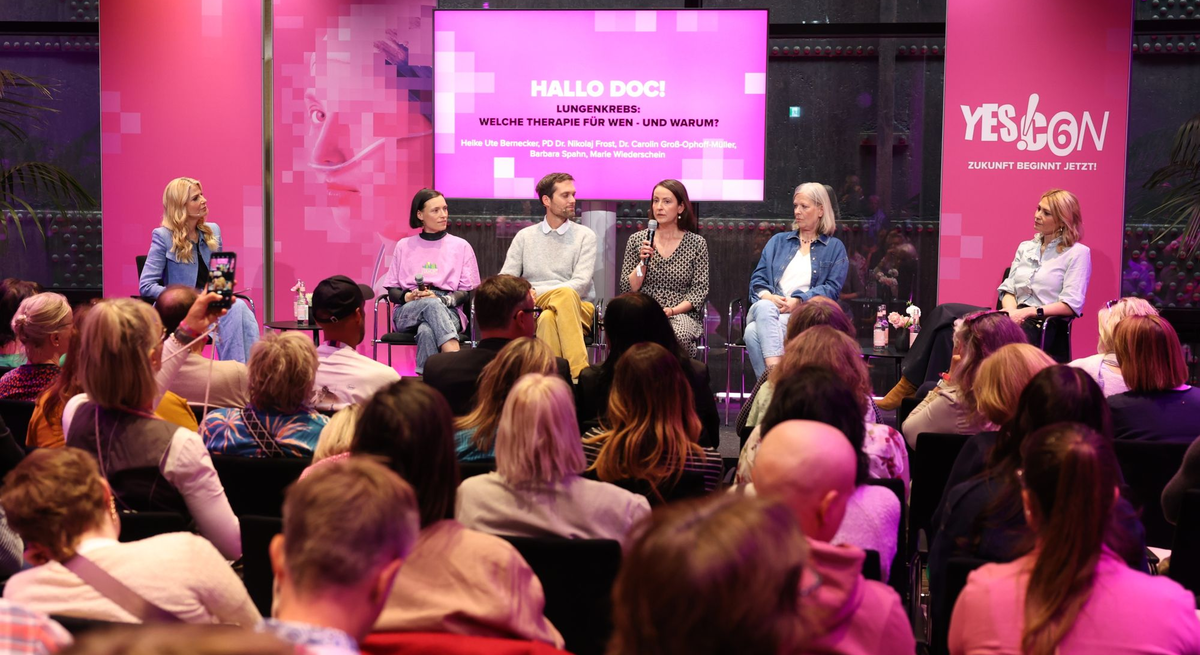 YES!CON: Fünf Personen diskutieren auf einer Bühne in heller Kleidung, hinter ihnen rosa Werbebanner, davor sitzt ein aufmerksam lauschendes Publikum. | © Jörg Carstensen