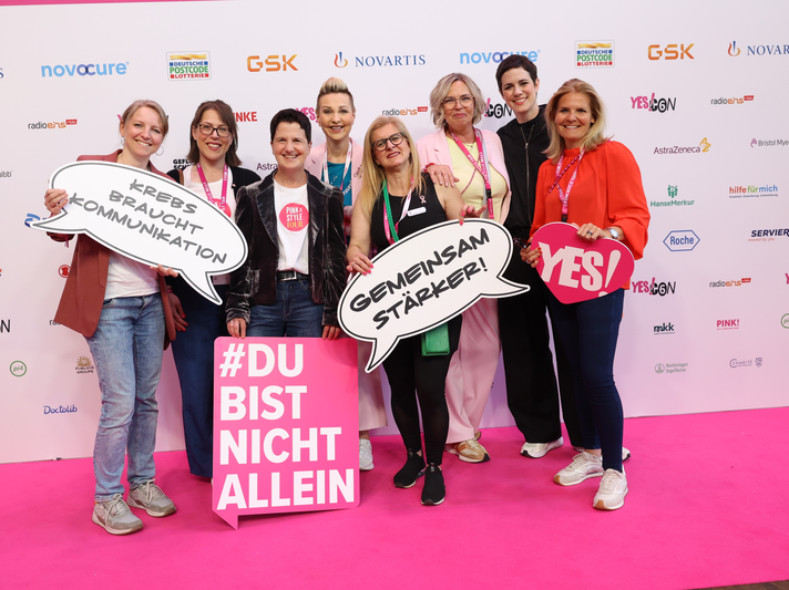 YES!CON: Sechs Personen stehen auf pinkem Teppich vor einer Sponsorenwand, halten Schilder mit motivierenden Botschaften zu Krebs. | © Jörg Carstensen