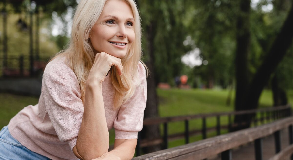 Das Foto zeigt eine lächelnde blonde Frau, die entspannt im Freien sitzt und sich an eine hölzerne Brüstung lehnt. Sie trägt ein rosa Oberteil und eine blaue Jeans. Ihr Blick ist nachdenklich und sie scheint etwas in der Ferne zu betrachten. Der Hintergrund ist unscharf, mit Bäumen und Grünflächen, was auf einen Park oder eine ähnliche natürliche Umgebung hindeutet. Die entspannte Pose und das freundliche Lächeln der Frau vermitteln eine ruhige und zufriedene Ausstrahlung.