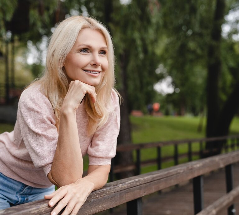 Das Foto zeigt eine lächelnde blonde Frau, die entspannt im Freien sitzt und sich an eine hölzerne Brüstung lehnt. Sie trägt ein rosa Oberteil und eine blaue Jeans. Ihr Blick ist nachdenklich und sie scheint etwas in der Ferne zu betrachten. Der Hintergrund ist unscharf, mit Bäumen und Grünflächen, was auf einen Park oder eine ähnliche natürliche Umgebung hindeutet. Die entspannte Pose und das freundliche Lächeln der Frau vermitteln eine ruhige und zufriedene Ausstrahlung. | © Adobe Stock/ InsideCreativeHouse