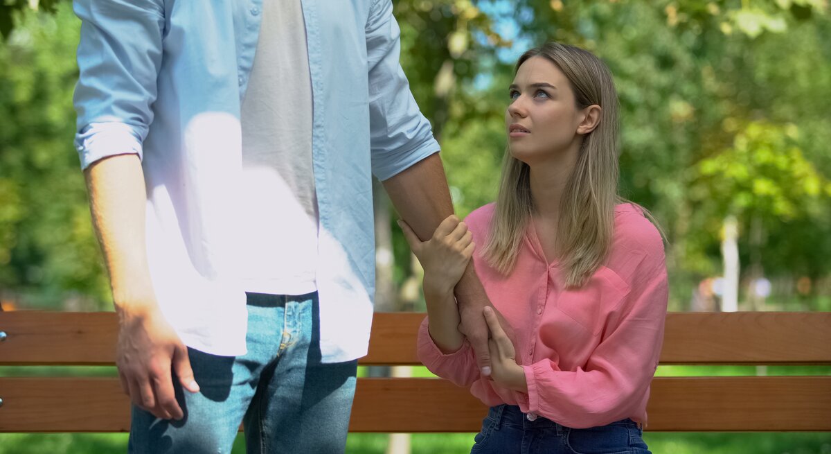 Mann lässt verärgerte Freundin allein auf Bank im Park zurück. Frau hält den Arm des Mannes fest.