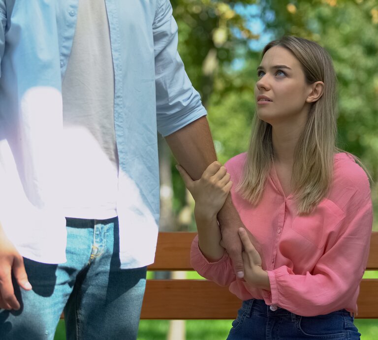 Mann lässt verärgerte Freundin allein auf Bank im Park zurück. Frau hält den Arm des Mannes fest. | © Motortion via Getty Images