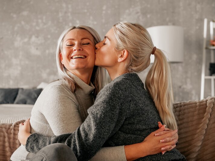 Eine ältere und eine jüngere Frau sitzen auf einer Couch und umarmen sich. Sie jüngere Frau gibt der älteren einen Kuss auf die Wange. | © Adobe Stock/ Vita