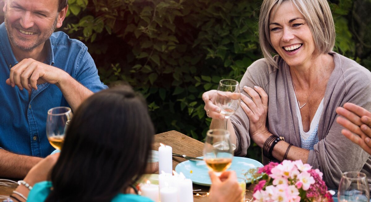 Eine Frau sitzt an einer Tafel bei einer Gartenparty und unterhält sich mit der Frau gegenüber. Die Runde sieht glücklich und entspannt aus.