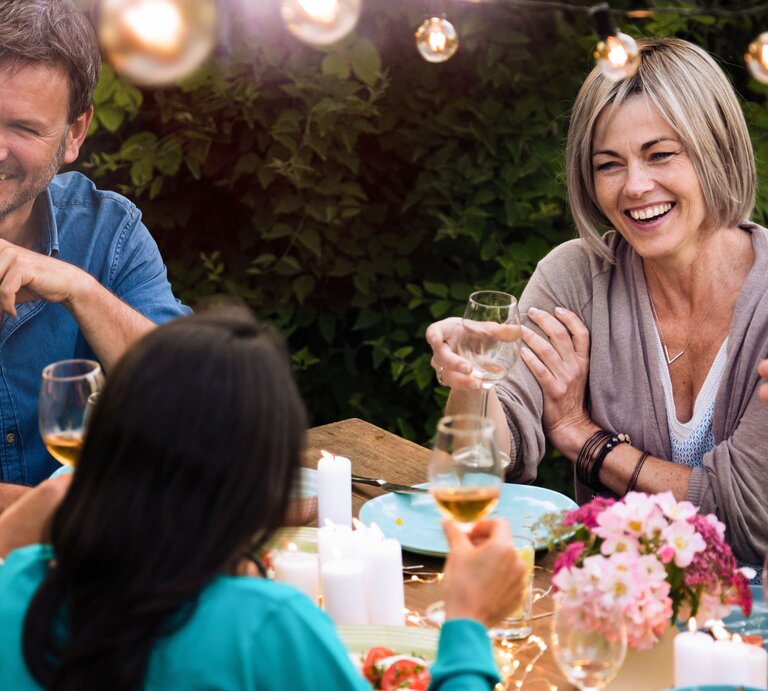 Eine Frau sitzt an einer Tafel bei einer Gartenparty und unterhält sich mit der Frau gegenüber. Die Runde sieht glücklich und entspannt aus. | © Adobe Stock/ jackfrog