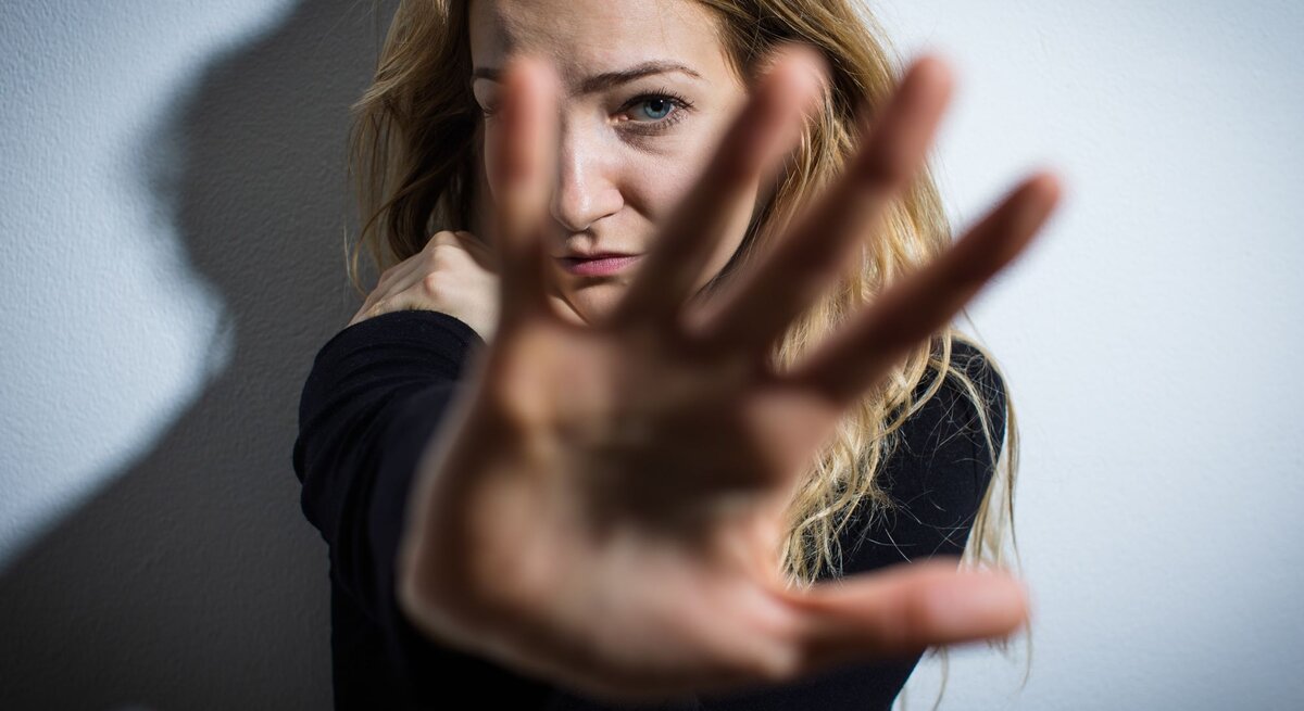 Das Foto zeigt eine junge Frau, die ihre Hand zur Kamera hin ausstreckt, als ob sie versucht, die Aufnahme zu blockieren oder sich zu schützen. Ihr Gesichtsausdruck wirkt besorgt oder ängstlich. Die Beleuchtung erzeugt einen dramatischen Kontrast und Schatten an der Wand hinter ihr, was die emotionale Intensität des Bildes verstärkt. Es könnte eine Darstellung von Abwehr, Privatsphäre oder einer emotionalen Grenze sein.