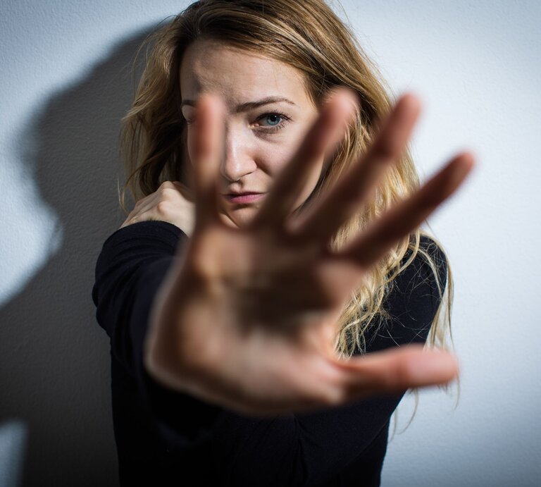 Das Foto zeigt eine junge Frau, die ihre Hand zur Kamera hin ausstreckt, als ob sie versucht, die Aufnahme zu blockieren oder sich zu schützen. Ihr Gesichtsausdruck wirkt besorgt oder ängstlich. Die Beleuchtung erzeugt einen dramatischen Kontrast und Schatten an der Wand hinter ihr, was die emotionale Intensität des Bildes verstärkt. Es könnte eine Darstellung von Abwehr, Privatsphäre oder einer emotionalen Grenze sein. | © IMAGO / Panthermedia