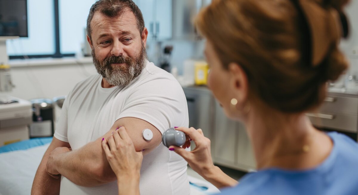 Älterer Mann mit grauem Bart lässt in Klinik von Ärztin im blauen Kittel seinen Glukosesensor am Oberarm scannen.