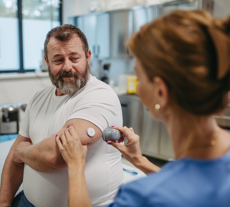 Älterer Mann mit grauem Bart lässt in Klinik von Ärztin im blauen Kittel seinen Glukosesensor am Oberarm scannen. | © Adobe Stock / Halfpoint