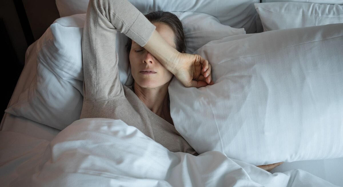 Eine Frau mit blassem Gesicht liegt erschöpft in einem weißen Bett, trägt einen beigefarbenen Pullover und hält eine Hand über die Augen