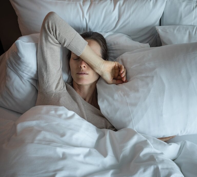Eine Frau mit blassem Gesicht liegt erschöpft in einem weißen Bett, trägt einen beigefarbenen Pullover und hält eine Hand über die Augen | © AdobeStock/ kieferpix