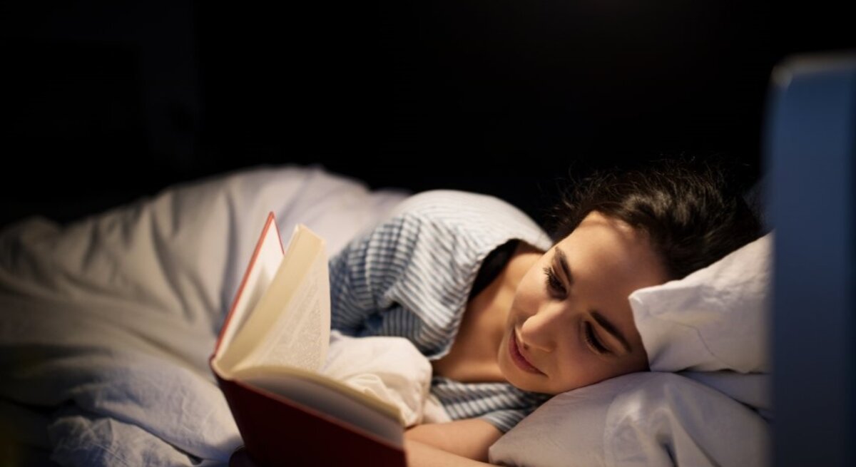 Junge Frau mit dunklen Haaren liest entspannt ein Buch im Bett, beleuchtet von einer warmen Nachttischlampe in dunkler Umgebung.