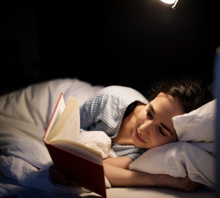 Junge Frau mit dunklen Haaren liest entspannt ein Buch im Bett, beleuchtet von einer warmen Nachttischlampe in dunkler Umgebung. | © Getty Images / Luis Alvarez