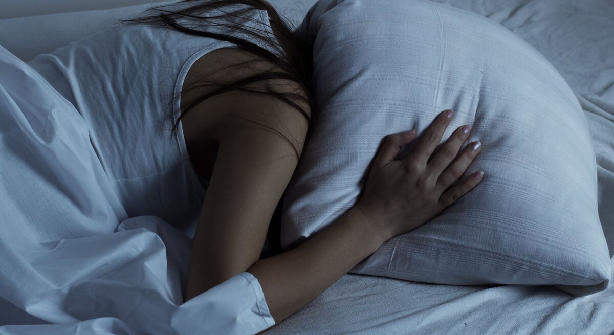 Eine junge Frau in weißem Nachthemd liegt seitlich im Bett, umarmt ein Kissen und wirkt nachdenklich, als ob sie von einem Gedankenkarussell geplagt wird.