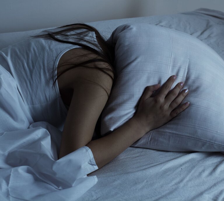 Eine junge Frau in weißem Nachthemd liegt seitlich im Bett, umarmt ein Kissen und wirkt nachdenklich, als ob sie von einem Gedankenkarussell geplagt wird. | © AdobeStock/mitarart