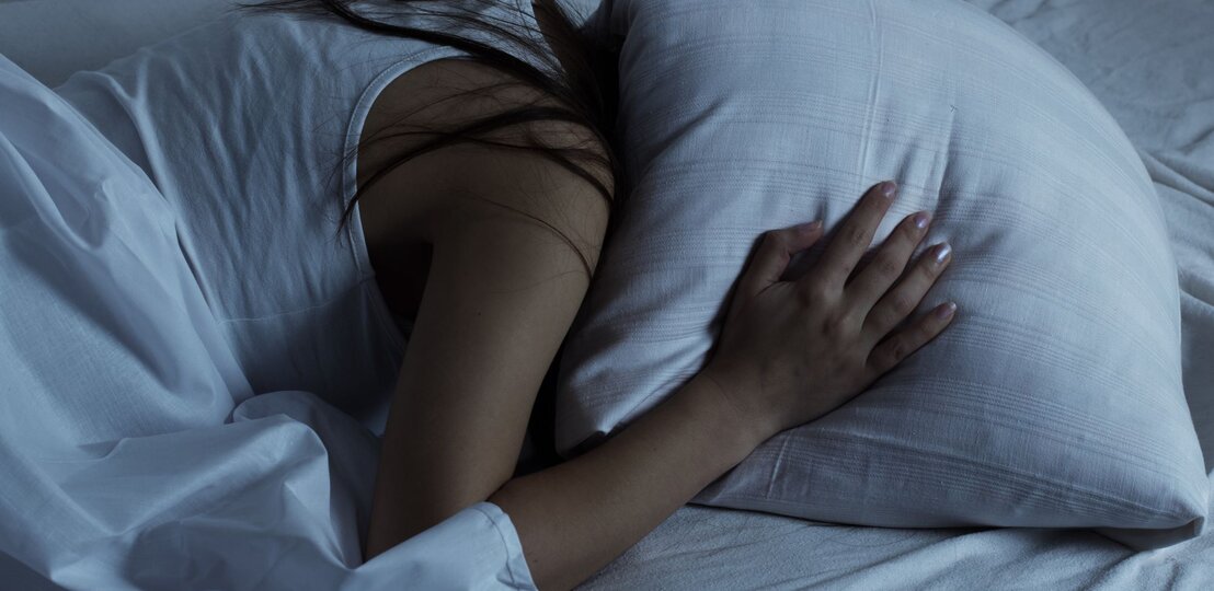 Eine junge Frau in weißem Nachthemd liegt seitlich im Bett, umarmt ein Kissen und wirkt nachdenklich, als ob sie von einem Gedankenkarussell geplagt wird. | © AdobeStock/mitarart