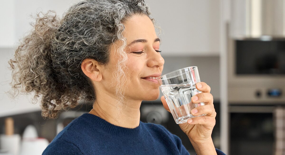 Eine Frau mit grauen Locken und blauem Pullover trinkt lächelnd ein Glas Wasser in einer modernen Küche.