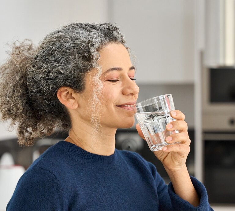 Eine Frau mit grauen Locken und blauem Pullover trinkt lächelnd ein Glas Wasser in einer modernen Küche. | © stock.adobe.com – insta_photos 
