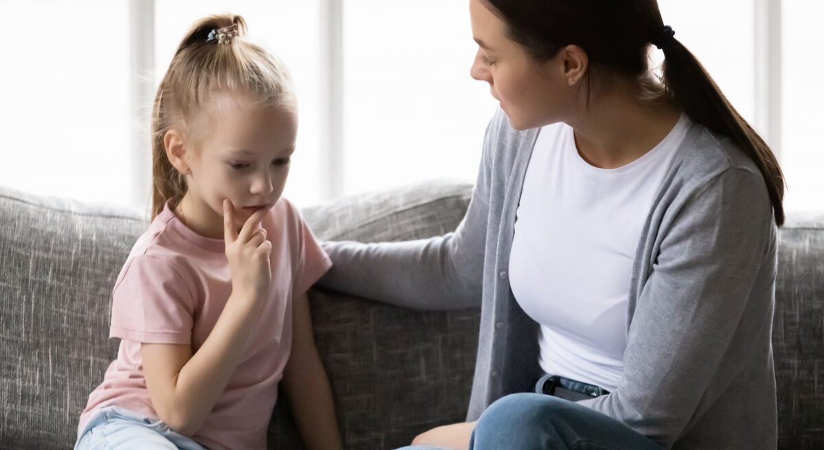 Eine junge Frau in grauem Cardigan sitzt beruhigend neben einem nachdenklichen Mädchen mit blondem Pferdeschwanz auf einem Sofa.