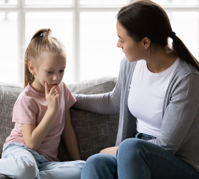 Eine junge Frau in grauem Cardigan sitzt beruhigend neben einem nachdenklichen Mädchen mit blondem Pferdeschwanz auf einem Sofa. | © Adobe Stock/fizkes