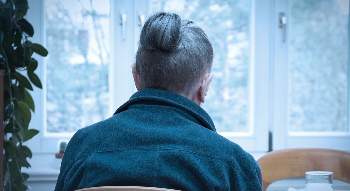 Ältere Frau, die alleine an einem Tisch sitzt, mit dem Rücken zur Kamera. Sie trägt eine blaue Jacke, und ihre grauen Haare sind zu einem Knoten hochgesteckt.