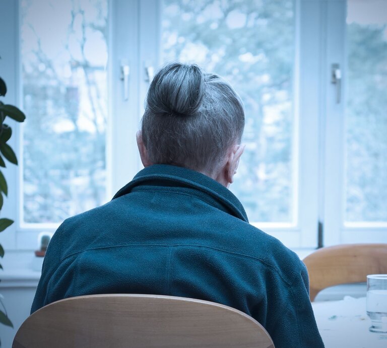 Ältere Frau, die alleine an einem Tisch sitzt, mit dem Rücken zur Kamera. Sie trägt eine blaue Jacke, und ihre grauen Haare sind zu einem Knoten hochgesteckt. | © Adobe Stock / agenturfotografin