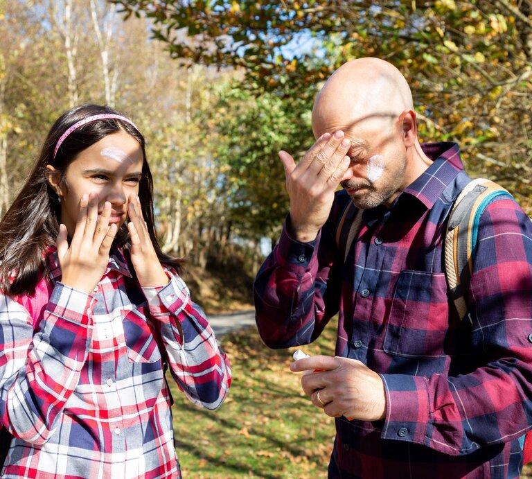 Ein Mann und ein junges Mädchen mit karierter Kleidung tragen Sonnencreme auf, während sie auf einem herbstlichen Waldweg stehen. | © Adobe Stock/asife