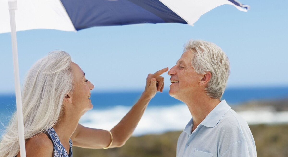 Lachende ältere Frau mit langen grauen Haaren trägt Sonnencreme auf die Nase eines ebenfalls lachenden Mannes am Strand auf.
