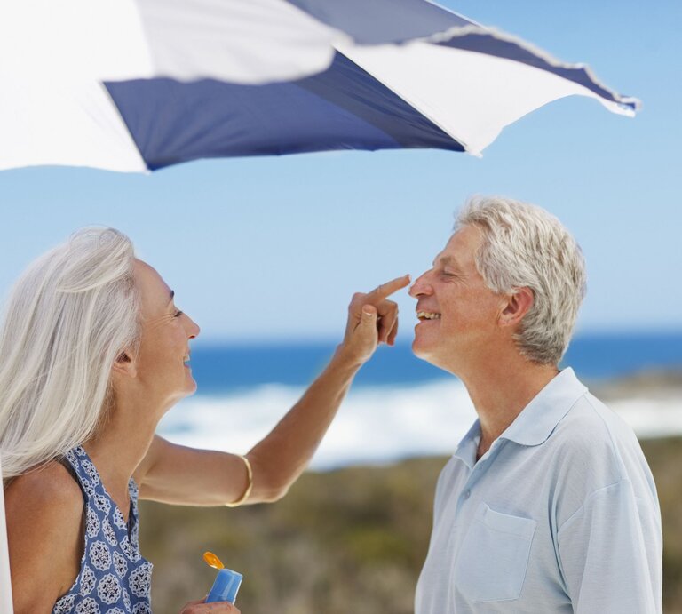 Lachende ältere Frau mit langen grauen Haaren trägt Sonnencreme auf die Nase eines ebenfalls lachenden Mannes am Strand auf. | © Getty Images/Tom Merton