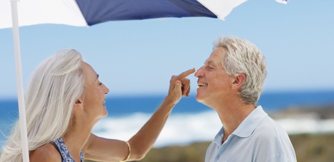 Lachende ältere Frau mit langen grauen Haaren trägt Sonnencreme auf die Nase eines ebenfalls lachenden Mannes am Strand auf. | © Getty Images/Tom Merton