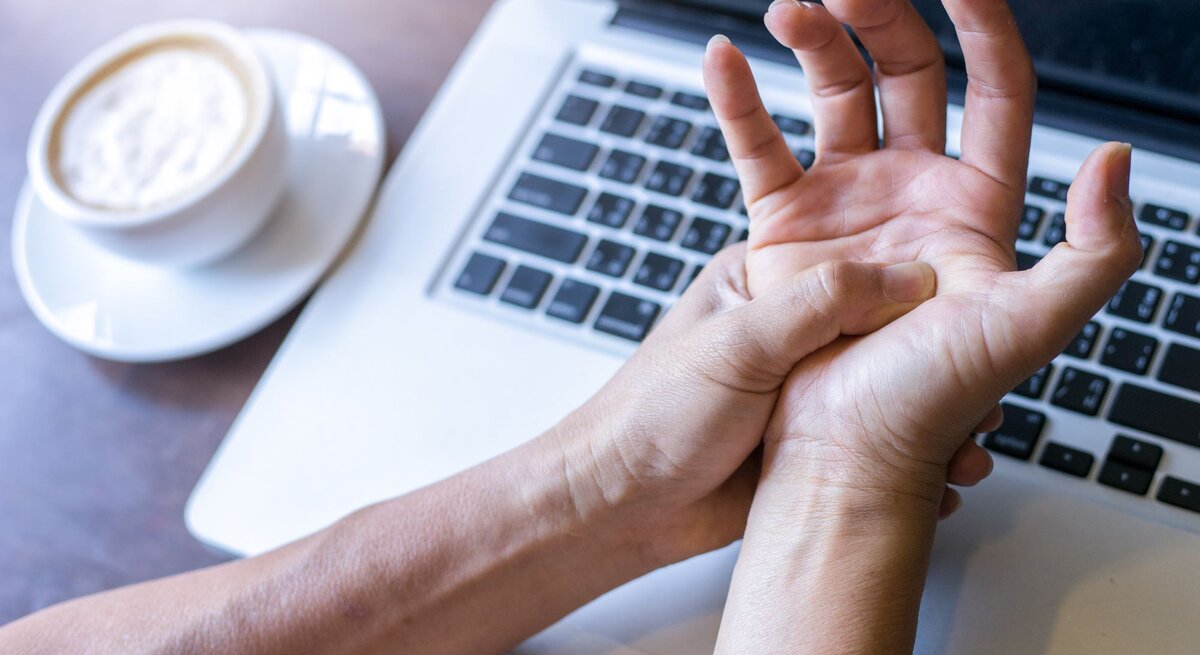 Eine Person hält sich vor einem Laptop und einer Tasse Kaffee den schmerzenden Handgelenk.