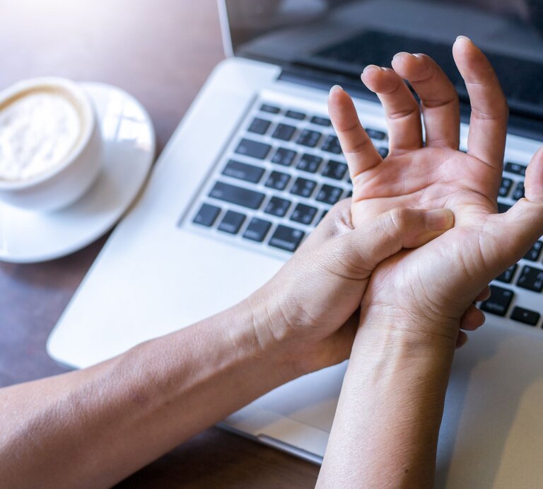 Eine Person hält sich vor einem Laptop und einer Tasse Kaffee den schmerzenden Handgelenk. | © GettyImages/krisanapong detraphiphat