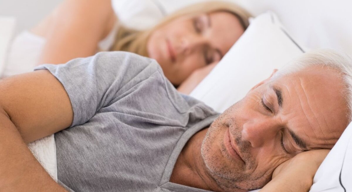 Das Bild zeigt ein älteres Paar, das friedlich im Bett schläft. Der Mann liegt im Vordergrund und scheint tief zu schlafen, mit einer Hand unter seinem Kopf und geschlossenen Augen. Er hat kurzes graues Haar und trägt ein graues T-Shirt. Die Frau liegt im Hintergrund und ist ebenfalls schlafend dargestellt, mit dem Kopf auf einem Kissen und dem Gesicht teils vom Bettrahmen verdeckt. Sie hat blondes Haar und trägt ein weißes Oberteil. Die Szene wirkt ruhig und entspannt, mit weicher Beleuchtung, die eine gemütliche Atmosphäre schafft. Es ist ein intimes und privates Bild, das Ruhe und Komfort in einem häuslichen Umfeld darstellt.