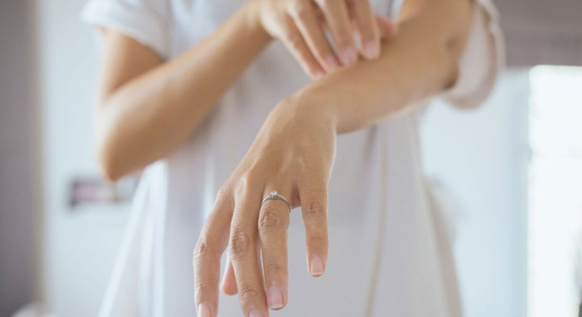  Das Bild zeigt eine Person, die ihren eigenen Unterarm betrachtet und mit der anderen Hand leicht darüber streicht oder kratzt. Das Individuum trägt ein weißes T-Shirt und hat einen Ring am Finger, was darauf hindeuten könnte, dass es sich um eine Frau handelt. Die Aufmerksamkeit auf den Arm und die Geste des Betrachtens oder Kratzens könnten auf Hautprobleme wie Nesselsucht hinweisen, bei der die Betroffenen oft einen Juckreiz oder eine Reizung verspüren. 
