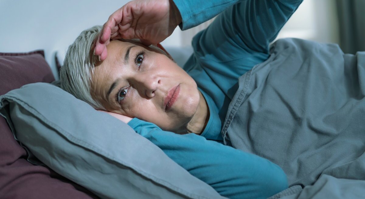 Das Bild zeigt eine ältere Frau mit kurzen grauen Haaren, die im Bett liegt und offenbar Schwierigkeiten beim Schlafen hat. Sie stützt ihren Kopf mit einer Hand, während sie nachdenklich oder besorgt nach oben schaut. Die Frau trägt ein türkisfarbenes Pyjamaoberteil und liegt unter einer grauen Bettdecke, die gut zu den lila Kissen passt. Das Bild könnte die Problematik der Schlafschwierigkeiten bei älteren Menschen illustrieren, die manchmal als senile Bettflucht bezeichnet werden.