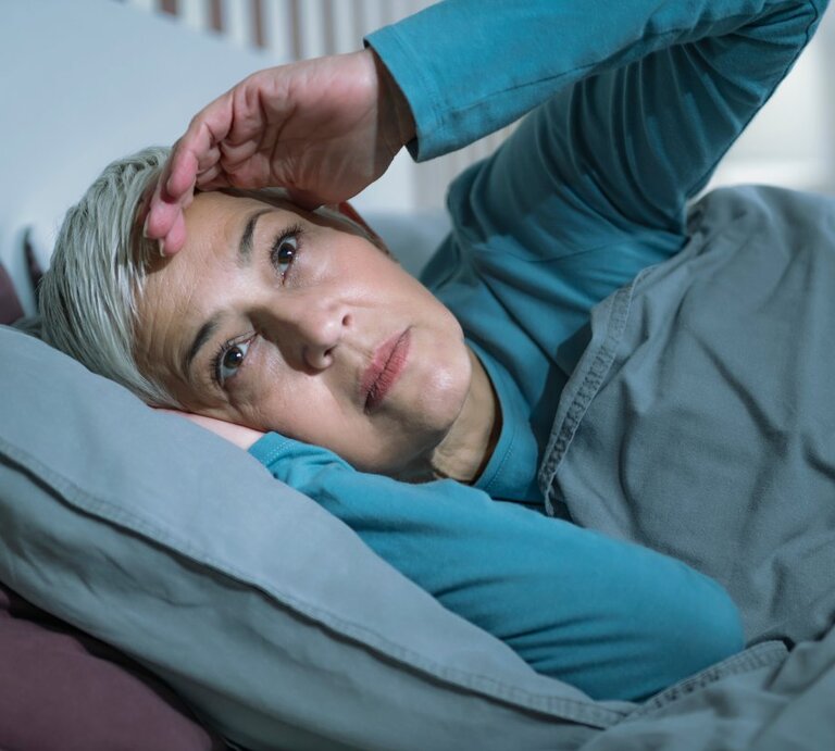 Das Bild zeigt eine ältere Frau mit kurzen grauen Haaren, die im Bett liegt und offenbar Schwierigkeiten beim Schlafen hat. Sie stützt ihren Kopf mit einer Hand, während sie nachdenklich oder besorgt nach oben schaut. Die Frau trägt ein türkisfarbenes Pyjamaoberteil und liegt unter einer grauen Bettdecke, die gut zu den lila Kissen passt. Das Bild könnte die Problematik der Schlafschwierigkeiten bei älteren Menschen illustrieren, die manchmal als senile Bettflucht bezeichnet werden. | © Getty Images/MICROGEN IMAGES/SCIENCE PHOTO LIBRARY