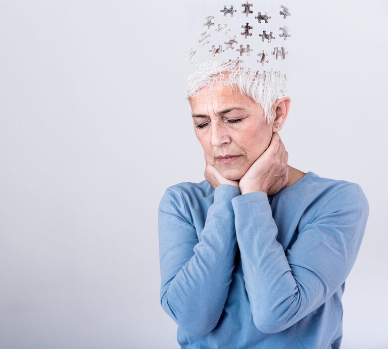 Grauhaarige Frau im blauen Oberteil hält nachdenklich ihren Kopf, mit Puzzleteilen, die als Zeichen des Gedächtnisverlusts entweichen. | © AdobeStock/Graphicroyalty