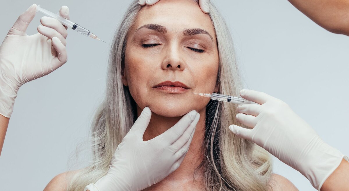 Das Bild zeigt eine Frau mittleren Alters, die eine kosmetische Behandlung erhält. Zwei Personen in medizinischen Handschuhen führen eine Injektionsbehandlung durch, wobei eine Person eine Spritze nahe der Stirn der Frau hält und die andere eine Spritze an ihrer Wange ansetzt. Solche Bilder sind typisch für ästhetische Eingriffe wie Botox- oder Filler-Behandlungen, die zur Minderung von Falten und zur Hautverjüngung eingesetzt werden. Die Frau hat geschlossene Augen und wirkt entspannt, was darauf hindeuten könnte, dass sie sich in einem professionellen kosmetischen oder medizinischen Umfeld befindet.