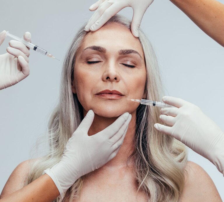 Das Bild zeigt eine Frau mittleren Alters, die eine kosmetische Behandlung erhält. Zwei Personen in medizinischen Handschuhen führen eine Injektionsbehandlung durch, wobei eine Person eine Spritze nahe der Stirn der Frau hält und die andere eine Spritze an ihrer Wange ansetzt. Solche Bilder sind typisch für ästhetische Eingriffe wie Botox- oder Filler-Behandlungen, die zur Minderung von Falten und zur Hautverjüngung eingesetzt werden. Die Frau hat geschlossene Augen und wirkt entspannt, was darauf hindeuten könnte, dass sie sich in einem professionellen kosmetischen oder medizinischen Umfeld befindet. | © Adobe Stock/ Jacob Lund